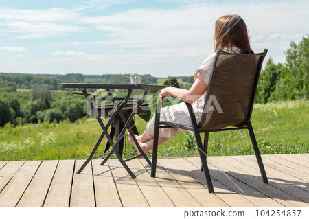 Woman relaxing on terrace, sitting in chair at table, enjoying view on nature landscape, forest valley on summer holiday Woman relaxing on terrace, sitting in chair at table, enjoying view on nature landscape, forest valley on summer holiday 104254857
