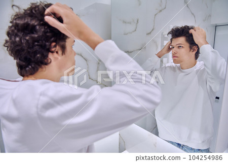 Young male looking at himself in bathroom mirror, touching curly hairstyle 104254986