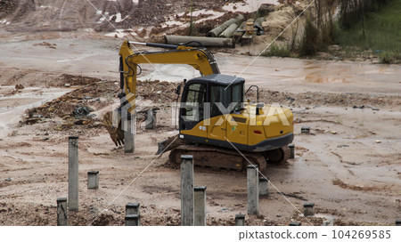 An excavator is working to insert stake or concrete foundation pillars into the ground on a construction site project. An excavator is working to insert stake or concrete foundation pillars into the ground on a construction site project. 104269585