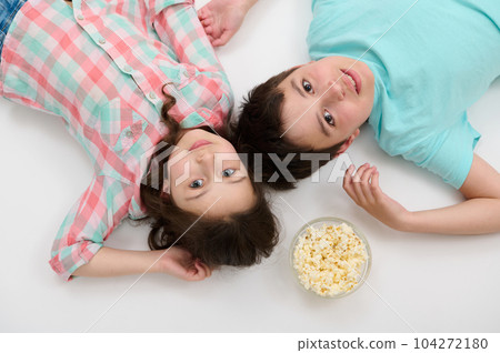 View from above of Caucasian adorable kids, beautiful little girl in checkered shirt and handsome preteen boy in blue t-shirt, lying on white background with a bowl of popcorn, looking at camera View from above of Caucasian adorable kids, beautiful little girl in checkered shirt and handsome preteen boy in blue t-shirt, lying on white background with a bowl of popcorn, looking at camera 104272180