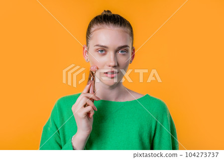 Young woman doing face massage with rose quartz stone roller. yellow background 104273737