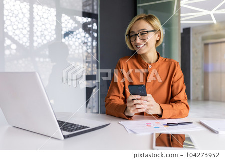 Young beautiful and successful businesswoman inside office at workplace with phone in hands, female employee smiling satisfied with results of achievement at work, blonde woman using app smartphone. Young beautiful and successful businesswoman inside office at workplace with phone in hands, female employee smiling satisfied with results of achievement at work, blonde woman using app smartphone. 104273952