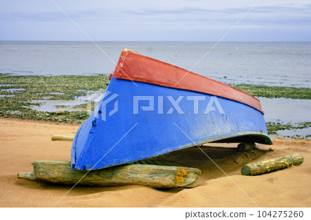 A blue-red boat lies upside down on the sandy seashore. 104275260