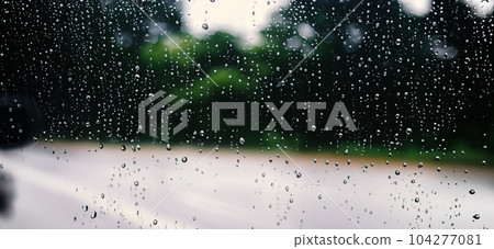 Rain shower on car windshield or car window and blurry road in background. Driving in rainy season. Rain drops on car mirror. Traffic road in evening rain. Drizzle raining decreases driving visibility 104277081