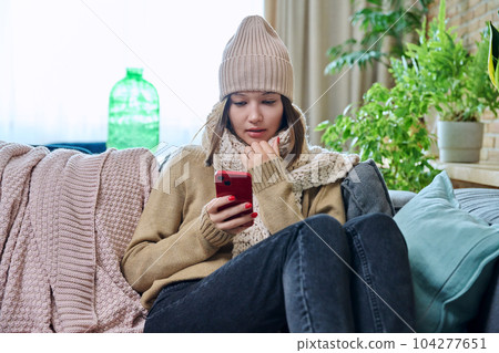 Young woman in sweater hat sitting at home, using smartphone Young woman in sweater hat sitting at home, using smartphone 104277651