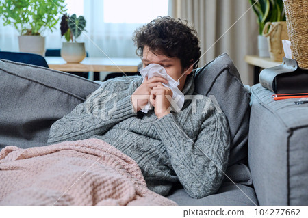 Sick young guy sneezing into nasal tissue, sitting on couch at home Sick young guy sneezing into nasal tissue, sitting on couch at home 104277662