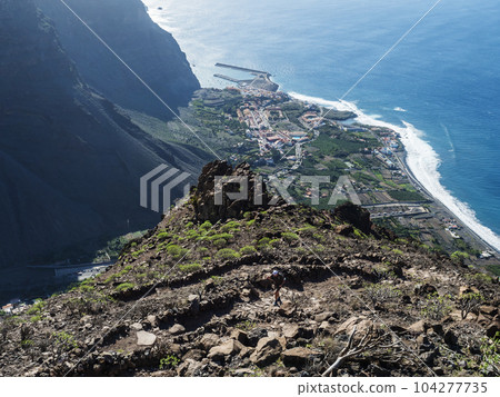 Aerial view of village resort Vueltas with port, Seen from top of La Merica mountain cliffs, Camino La Merica hiking trail. Valle Gran Rey, La Gomera, Canary Islands. Copy space. Aerial view of village resort Vueltas with port, Seen from top of La Merica mountain cliffs, Camino La Merica hiking trail. Valle Gran Rey, La Gomera, Canary Islands. Copy space. 104277735