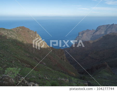 View of illuminated sharp rock and cliffs on top of La Merica mountain with cacti, pine and palm trees. Camino La Merica hiking trail. Arure, Valle Gran Rey , La Gomera, Canary Islands, Spain. View of illuminated sharp rock and cliffs on top of La Merica mountain with cacti, pine and palm trees. Camino La Merica hiking trail. Arure, Valle Gran Rey , La Gomera, Canary Islands, Spain. 104277744