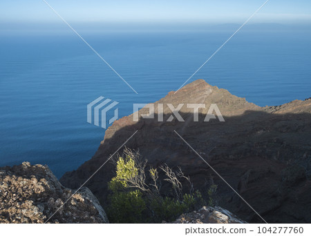 View of illuminated sharp rock and cliff on top of La Merica mountain. Camino La Merica hiking trail. Arure, Valle Gran Rey , La Gomera, Canary Islands, Spain. View of illuminated sharp rock and cliff on top of La Merica mountain. Camino La Merica hiking trail. Arure, Valle Gran Rey , La Gomera, Canary Islands, Spain. 104277760