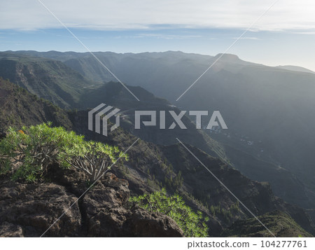 View from green plateau on top of La Merica mountain on valley with winding roads and hills in fog haze. Camino La Merica hiking trail. Valle Gran Rey, La Gomera, Canary Islands. Backlight, copy space View from green plateau on top of La Merica mountain on valley with winding roads and hills in fog haze. Camino La Merica hiking trail. Valle Gran Rey, La Gomera, Canary Islands. Backlight, copy space 104277761
