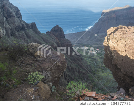 Aerial view of village Taguluche in green valley with palm trees, cacti and sharp cliffs of La Merica mountain. Seen from Camino La Merica hiking trail. Arure, Valle Gran Rey , La Gomera, Canary Aerial view of village Taguluche in green valley with palm trees, cacti and sharp cliffs of La Merica mountain. Seen from Camino La Merica hiking trail. Arure, Valle Gran Rey , La Gomera, Canary 104277781