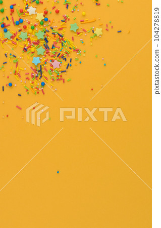 Decorative sprinkles border on yellow background. Multicolor Sugar confectionery powder in the corner. Scattered sweet sugar sticks, stars and balls. Copy space. Decorative sprinkles border on yellow background. Multicolor Sugar confectionery powder in the corner. Scattered sweet sugar sticks, stars and balls. Copy space. 104278819