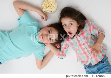 Happy children with popcorn bowl smile at camera, express surprise and amazement, lying on their backs on isolated white studio background. Kids. Childhood. Entertainment. Lifestyle. Leisure activity Happy children with popcorn bowl smile at camera, express surprise and amazement, lying on their backs on isolated white studio background. Kids. Childhood. Entertainment. Lifestyle. Leisure activity 104288800