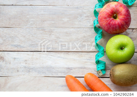 Healthy lifestyle concept with red apple. Weight loss or diet concept. Red apple and green measure tape,kiwi,carrot on wood table background Healthy lifestyle concept with red apple. Weight loss or diet concept. Red apple and green measure tape,kiwi,carrot on wood table background 104290284