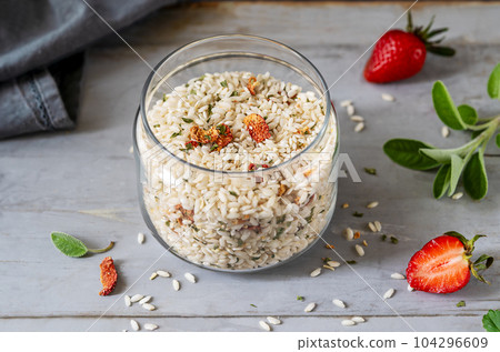 Risotto raw mix with strawberries and herbs in a glass container, fresh strawberries, sage, napkin over grey wooden background 104296609