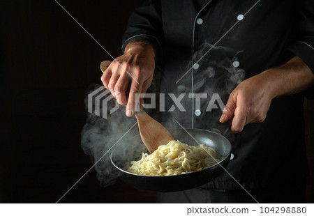The cook fries spaghetti in a hot pan. Space for advertising on a black background The cook fries spaghetti in a hot pan. Space for advertising on a black background 104298880