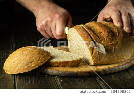 The cook hand with a knife slices bread on a kitchen cutting board. The concept of healthy food in the bakery 104298892