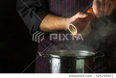 The cook adds ground pepper to a pot of boiling food. Retsoran kitchen cooking concept with advertising space on black background The cook adds ground pepper to a pot of boiling food. Retsoran kitchen cooking concept with advertising space on black background 104298901