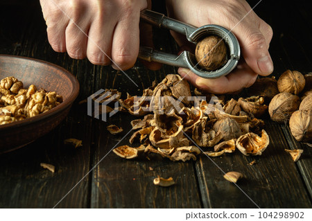 Peeling nuts with a nutcracker. Close-up of the chef's hands while working on the kitchen table. Heap of shelled walnuts in a bowl. Peeling nuts with a nutcracker. Close-up of the chef's hands while working on the kitchen table. Heap of shelled walnuts in a bowl. 104298902