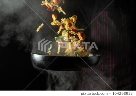Cook throws food on a hot pan. Thai street food pasta with spices and vegetables. Space for advertising on a dark background. Cook throws food on a hot pan. Thai street food pasta with spices and vegetables. Space for advertising on a dark background. 104298932