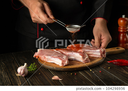 The cook adds paprika to raw meat ribs before grilling. Work environment on the kitchen table with spices and condiments The cook adds paprika to raw meat ribs before grilling. Work environment on the kitchen table with spices and condiments 104298936