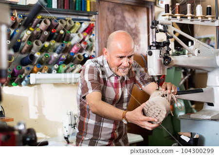 Mature workman sewing leather boots on stitch lathe 104300299