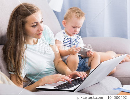 Satisfied woman is productively working behind laptop while kid playing on tablet 104300492