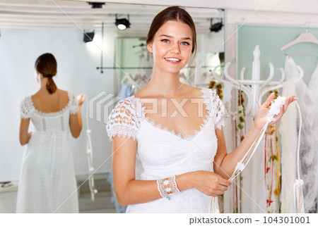 Young cheerful woman fitting luxury wedding dress at modern wedding salon 104301001