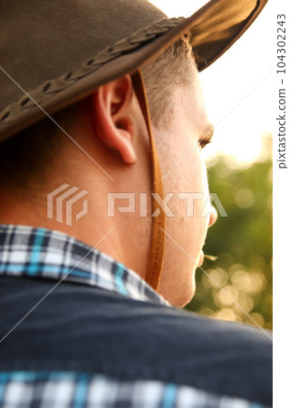 A young man wearing a cowboy hat gazes at the sky in the distance, capturing the essence of adventure and the spirit of the Wild West. The setting sun adds to the captivating atmosphere 104302243