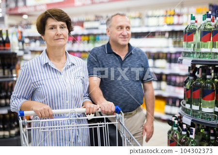 Mature family of retirees examines a bottle of vermouth in the alcohol section of a supermarket Mature family of retirees examines a bottle of vermouth in the alcohol section of a supermarket 104302373