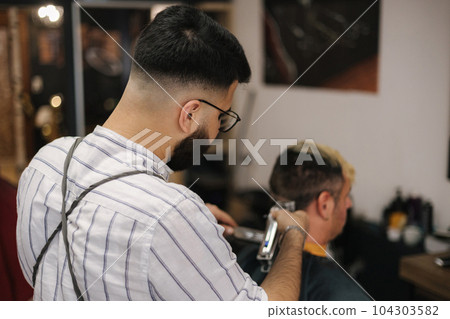 Bearded barber cutting hair of male customer. Hairstylist serving client at barber shop. Bearded barber cutting hair of male customer. Hairstylist serving client at barber shop. 104303582