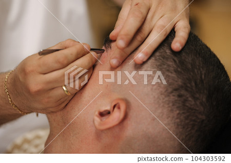 Close-up of barber man trimming eyebrows with tweezers for confident man Close-up of barber man trimming eyebrows with tweezers for confident man 104303592