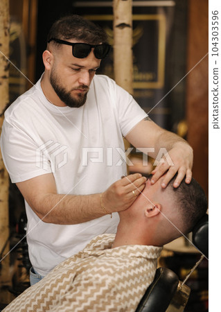 Side view of barber man trimming eyebrows with tweezers for confident man Side view of barber man trimming eyebrows with tweezers for confident man 104303596