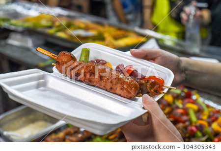 Hands holding plastic box with meat skewers kebab in vietnamese night market in food festival Hands holding plastic box with meat skewers kebab in vietnamese night market in food festival 104305343