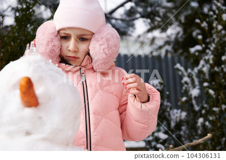 Caucasian adorable child 6 years old, lovely little girl in pink warm winter clothes and fluffy ear muffs, building a snowman in the snow covered backyard. Winter leisures activity. Christmas time 104306131