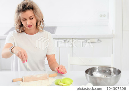 A young girl sits at a table in a bright white kitchen and prepares a lunch or a pastry dessert 104307806