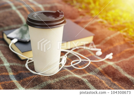 Cup of coffee, open book and smartphone with headphones on blanket in the park Cup of coffee, open book and smartphone with headphones on blanket in the park 104308673