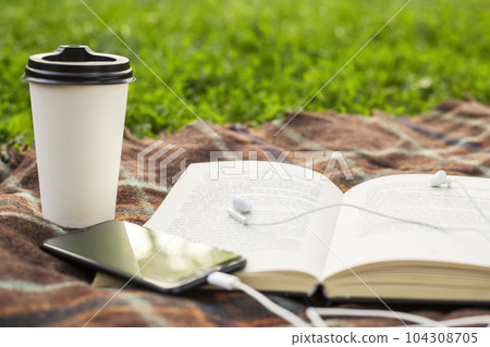 Cup of coffee, open book and smartphone with headphones on blanket in the park Cup of coffee, open book and smartphone with headphones on blanket in the park 104308705