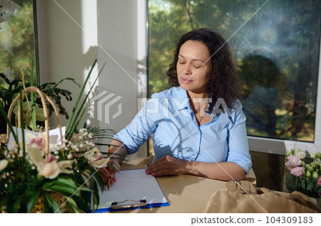 Inspired young female florist draws sketch with flower composition of a bouquet for Wedding Day, sitting in her office at floral store, surrounded by exotic flowers. People and small business concept Inspired young female florist draws sketch with flower composition of a bouquet for Wedding Day, sitting in her office at floral store, surrounded by exotic flowers. People and small business concept 104309183