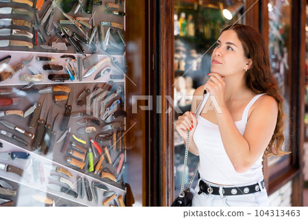Interested young woman viewing various knifes placed on display case Interested young woman viewing various knifes placed on display case 104313463