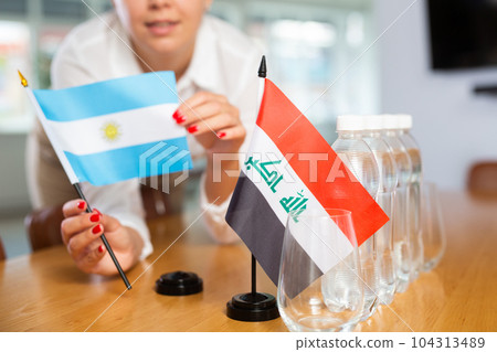 Woman secretary prepares an office for negotiations - she places flags of Iraq and Argentina table 104313489