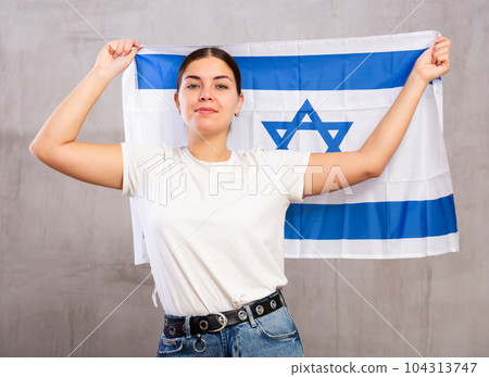 Portrait of serious young female holding national flag of Israel against gray wall 104313747
