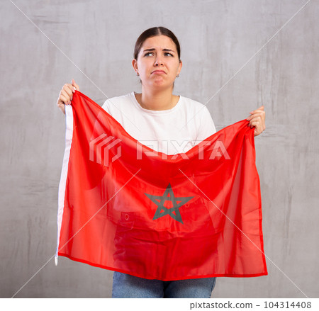 Stressed out sad young woman standing with flag of Morocco against gray wall Stressed out sad young woman standing with flag of Morocco against gray wall 104314408