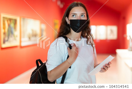 Young girl in a protective mask visits an exhibition of paintings in a museum 104314541
