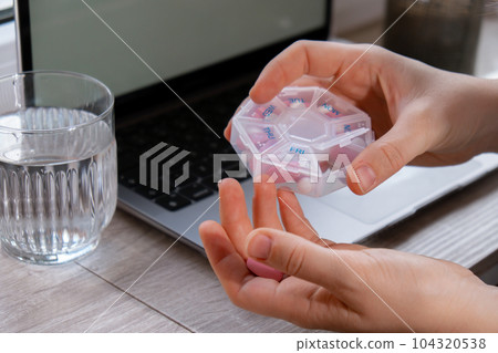 Woman taking Daily vitamins white working on laptop Organizer weekly shots Closeup of medical pill box with doses of tablets for daily take medicine with white pink drugs and capsules. Workplace 104320538
