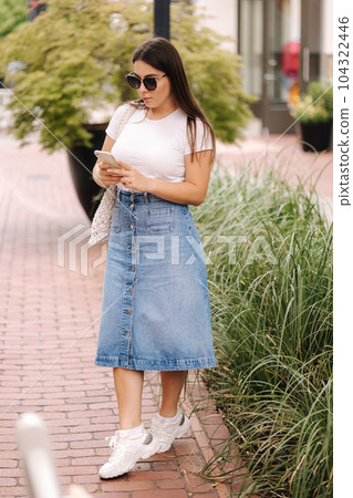 Beautiful young woman in denim skirt and white t-shirt, use phone outdoor during she waiting her friend 104322446