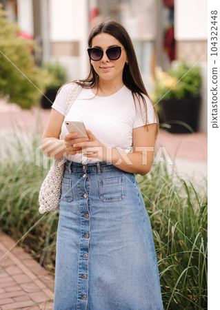 Pretty woman in denim skirt and white t-shirt using phone. Woman walking outdoors and looking for something in social networks 104322448