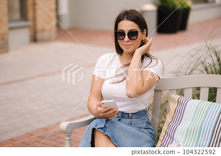 Portrait of beautiful woman sitting on the bench outdoor and using phone. Female in sunglasses Portrait of beautiful woman sitting on the bench outdoor and using phone. Female in sunglasses 104322592