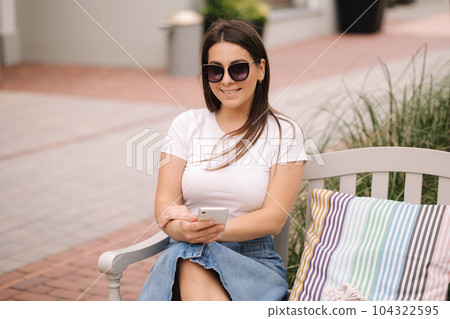 Portrait of beautiful woman sitting on the bench outdoor and using phone. Female in sunglasses Portrait of beautiful woman sitting on the bench outdoor and using phone. Female in sunglasses 104322595