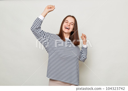 Extremely happy positive brown haired woman wearing striped shirt posing isolated over gray background standing with raised arms dancing celebrating holiday. 104323742